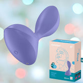 A lilac, curved personal massager designed for anal stimulation is displayed alongside its packaging box. The box showcases similar imagery and product branding of the Satisfyer Sweet Seal by Satisfyer. The softly blurred background features pastel-colored bokeh circles.